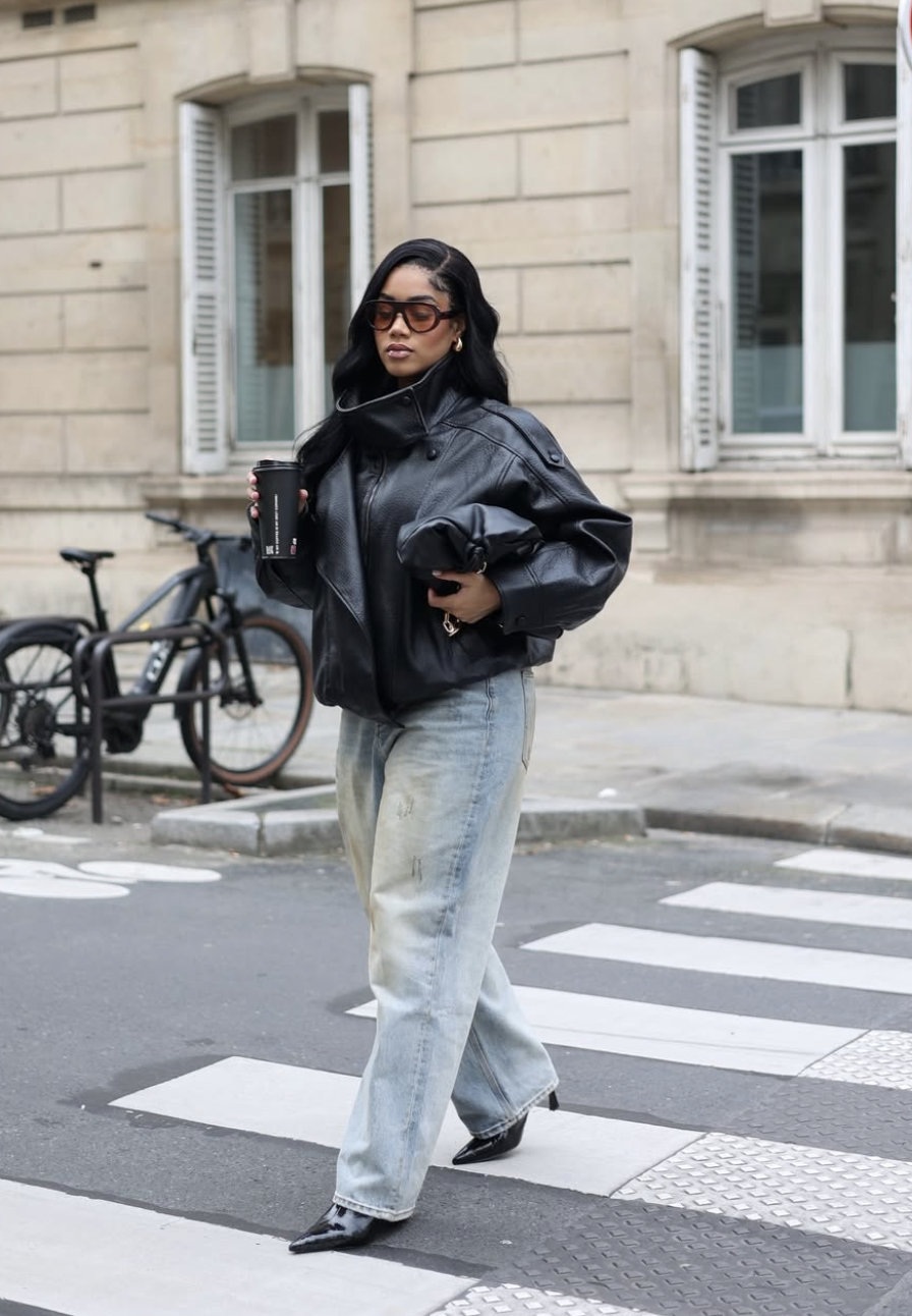 A woman crossing a street wearing light-wash wide-leg jeans, an oversized black leather jacket, and black pointed-toe boots.