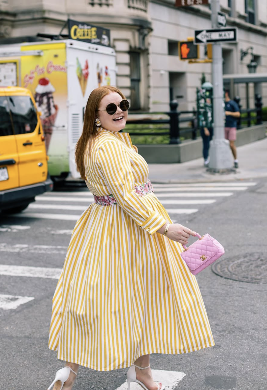 A street-style shot of a woman smiling while walking across a city street, wearing a vibrant yellow and white vertical-striped maxi dress styled with a floral woven waist belt, white ankle-strap heels, and a pink quilted mini handbag.