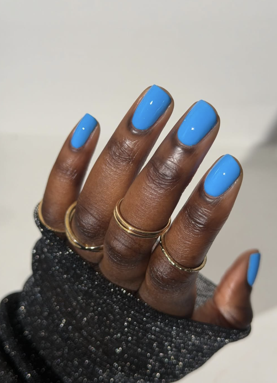 Close-up of a hand with long, rounded nails showcasing a vibrant, opaque electric blue manicure, styled with multiple gold rings and a black glittery mesh sleeve.