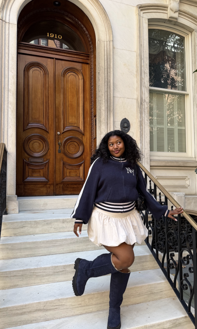Navy varsity-style sweater and white pleated skirt with tall dark UGG boots.