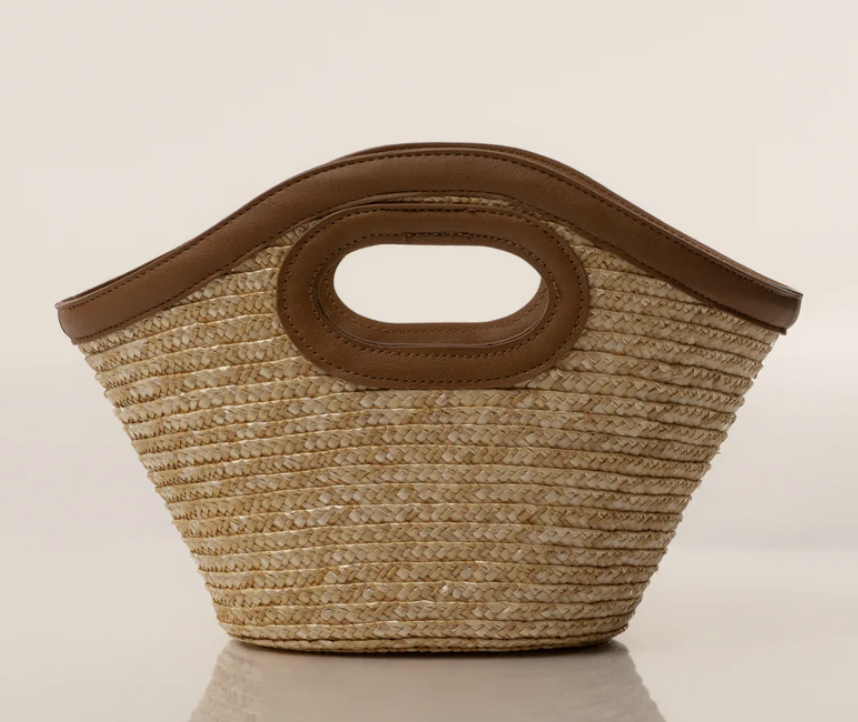 A structured tan woven straw tote bag with smooth chocolate brown leather handles, styled as a chic Mediterranean resort accessory.