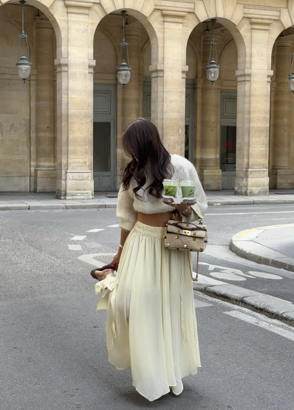 : A woman wearing a cream fuzzy cropped sweater and a matching flowing maxi skirt, carrying two matcha lattes and a studded handbag.