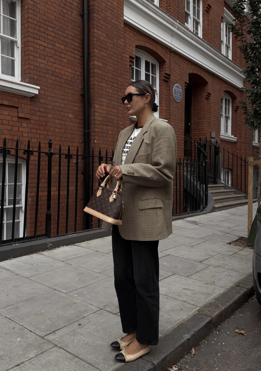 A woman wearing a white graphic t-shirt tucked into dark straight-leg trousers, styled with an oversized plaid blazer and loafers for a comfortable workwear look.