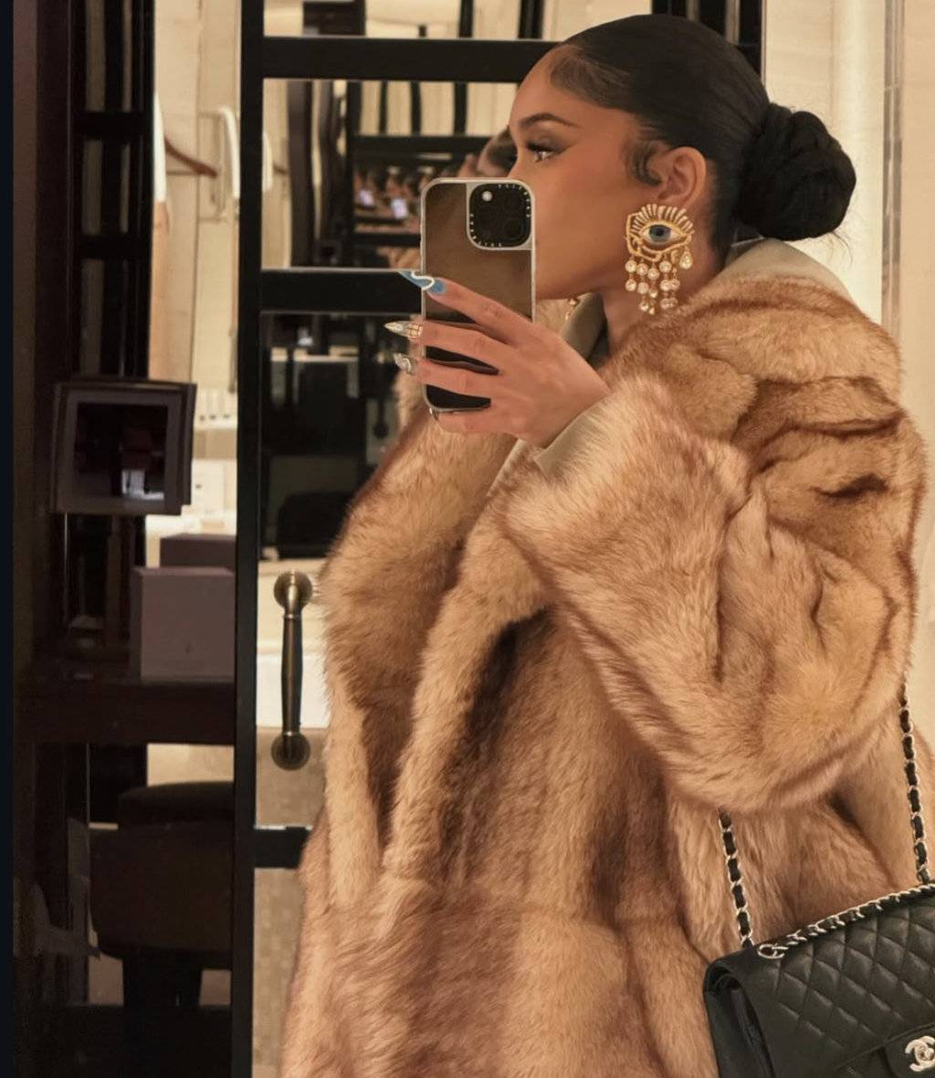 Saweetie with long, voluminous glam waves and a side part, wearing a fur coat and statement earrings, showing how side-swept volume adds definition to a round face shape.