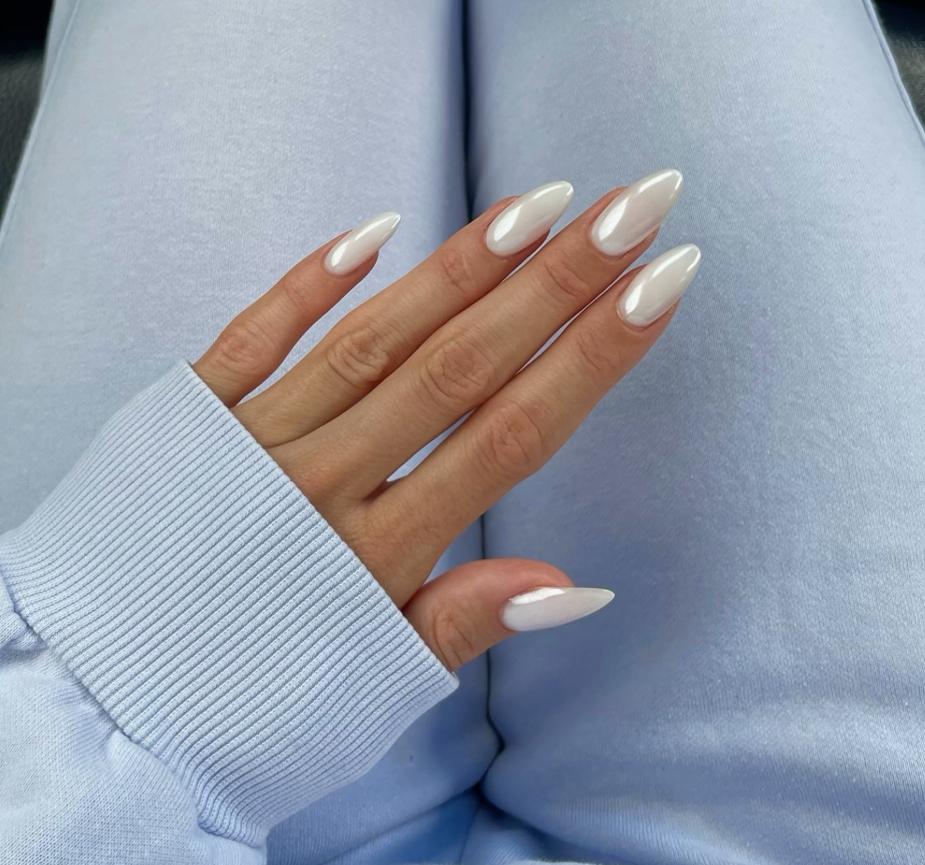 Close-up of a hand with long, almond-shaped nails showcasing a milky white chrome manicure with an iridescent, pearly finish, paired with a light blue ribbed sleeve.