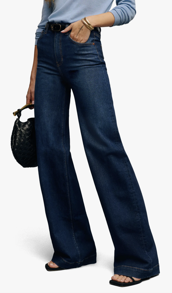A woman wearing dark wash wide-leg jeans and a light blue button-down shirt, showing a stylish casual work outfit that pairs perfectly with sneakers.