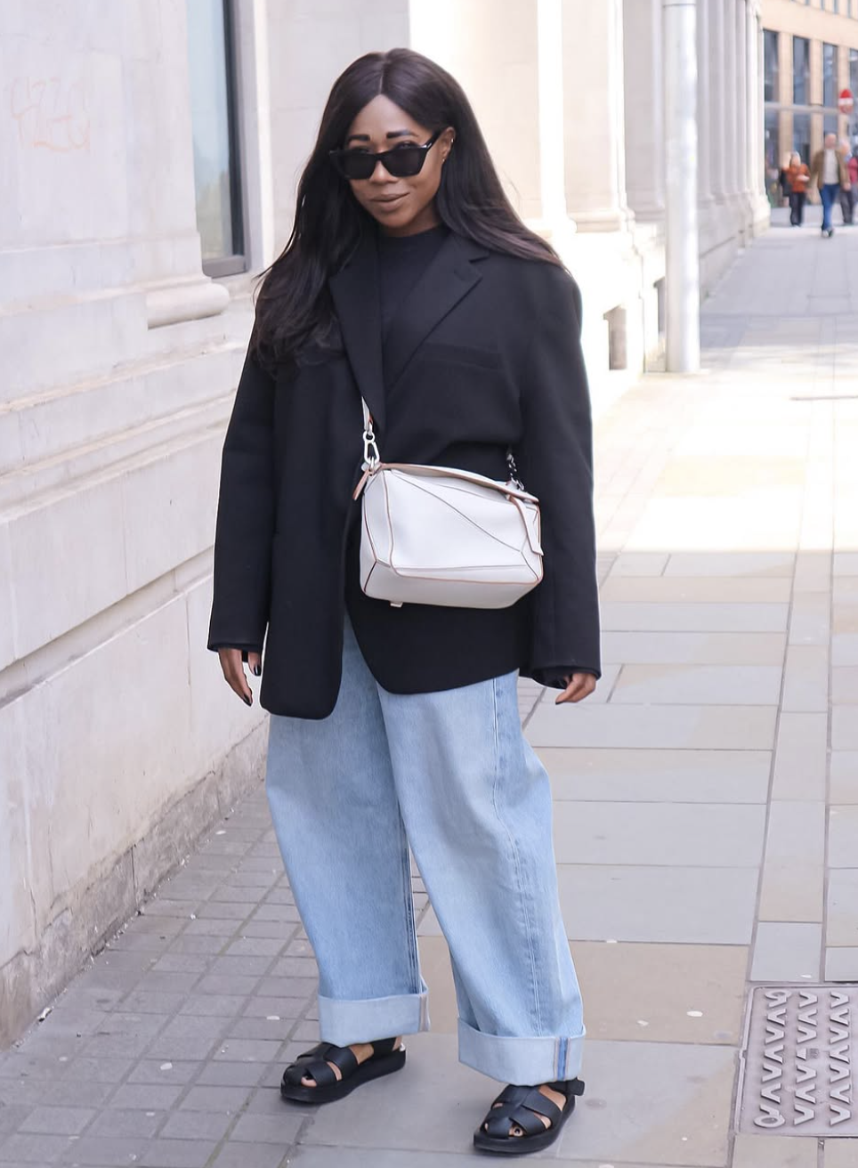 A woman wearing chunky black leather fisherman sandals with light wash slim-straight jeans, a black oversized blazer, and a white Loewe Puzzle bag.