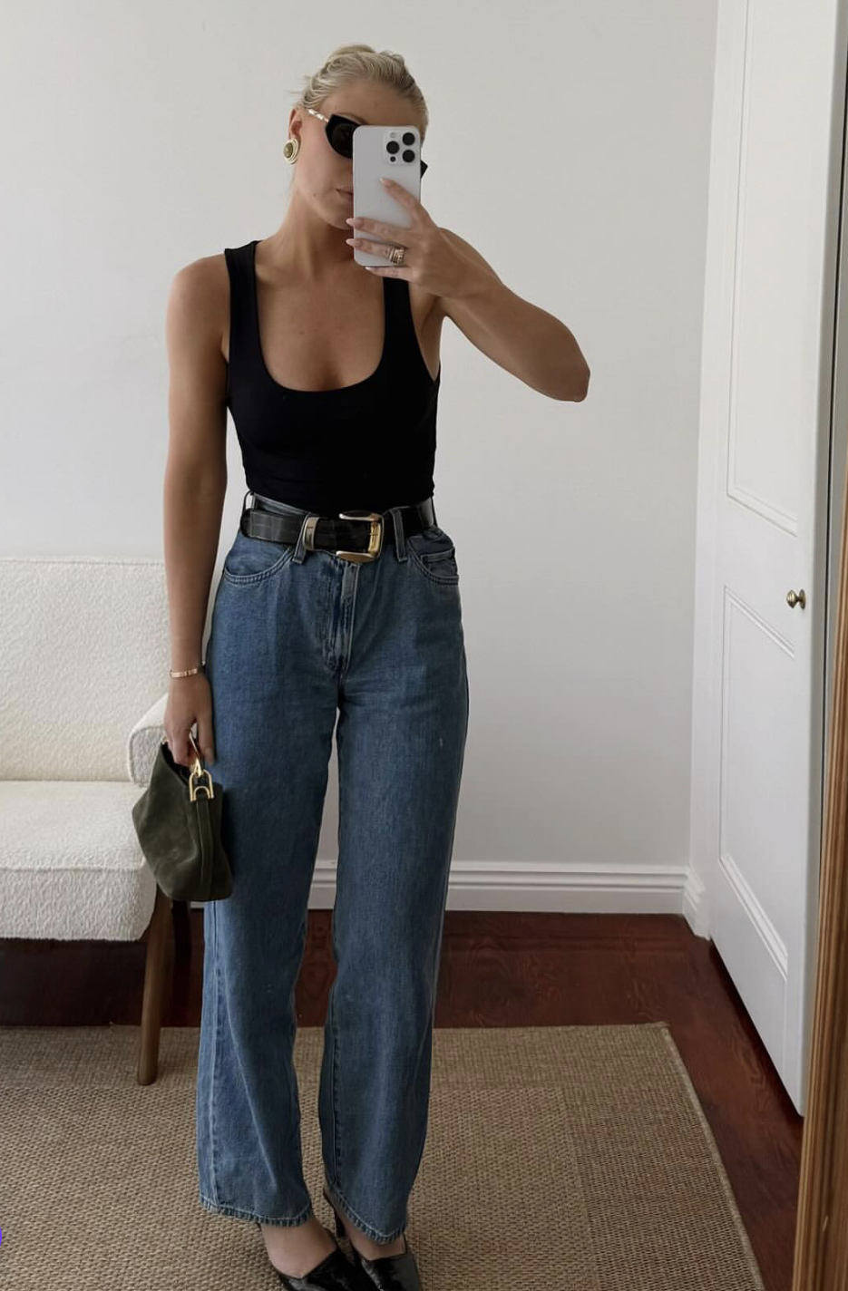 A woman wearing a black square-neck sleeveless bodysuit tucked into high-waisted blue jeans with a black belt, paired with black pointed-toe heels and a green handbag.