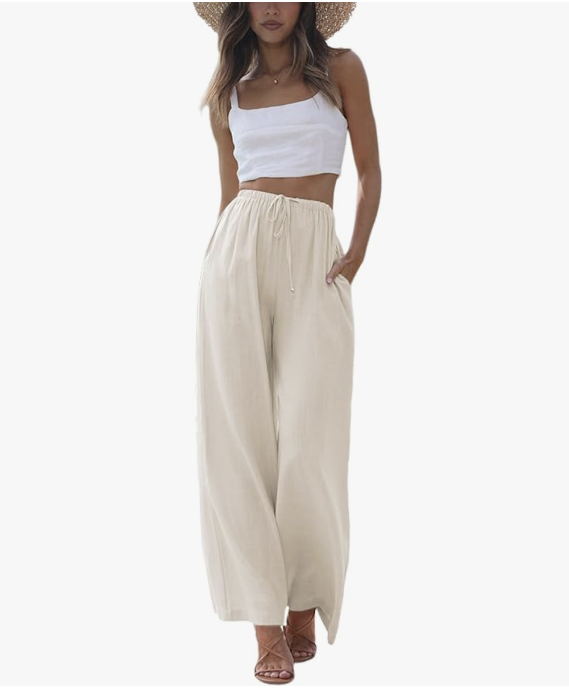 A woman wearing a white crop top and cream-colored high-waisted linen palazzo pants with a drawstring waist and side pockets, styled with a straw sun hat.