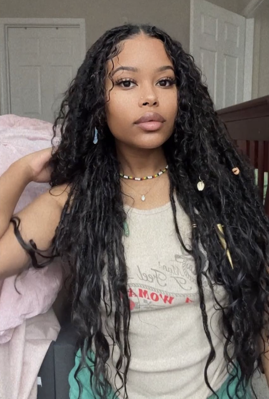 A woman with long, textured faux locs featuring curly ends and face-framing strands, wearing a colorful beaded necklace and a white tank top.
