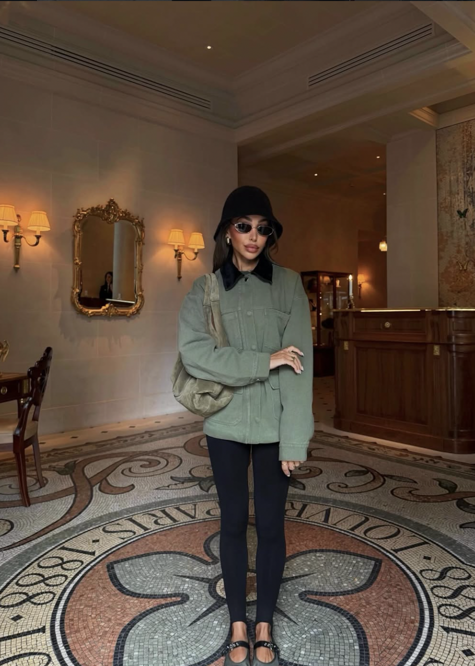 Woman wearing black stirrup leggings with a green button-up utility cardigan, a black bucket hat, and black leather ballet flats.