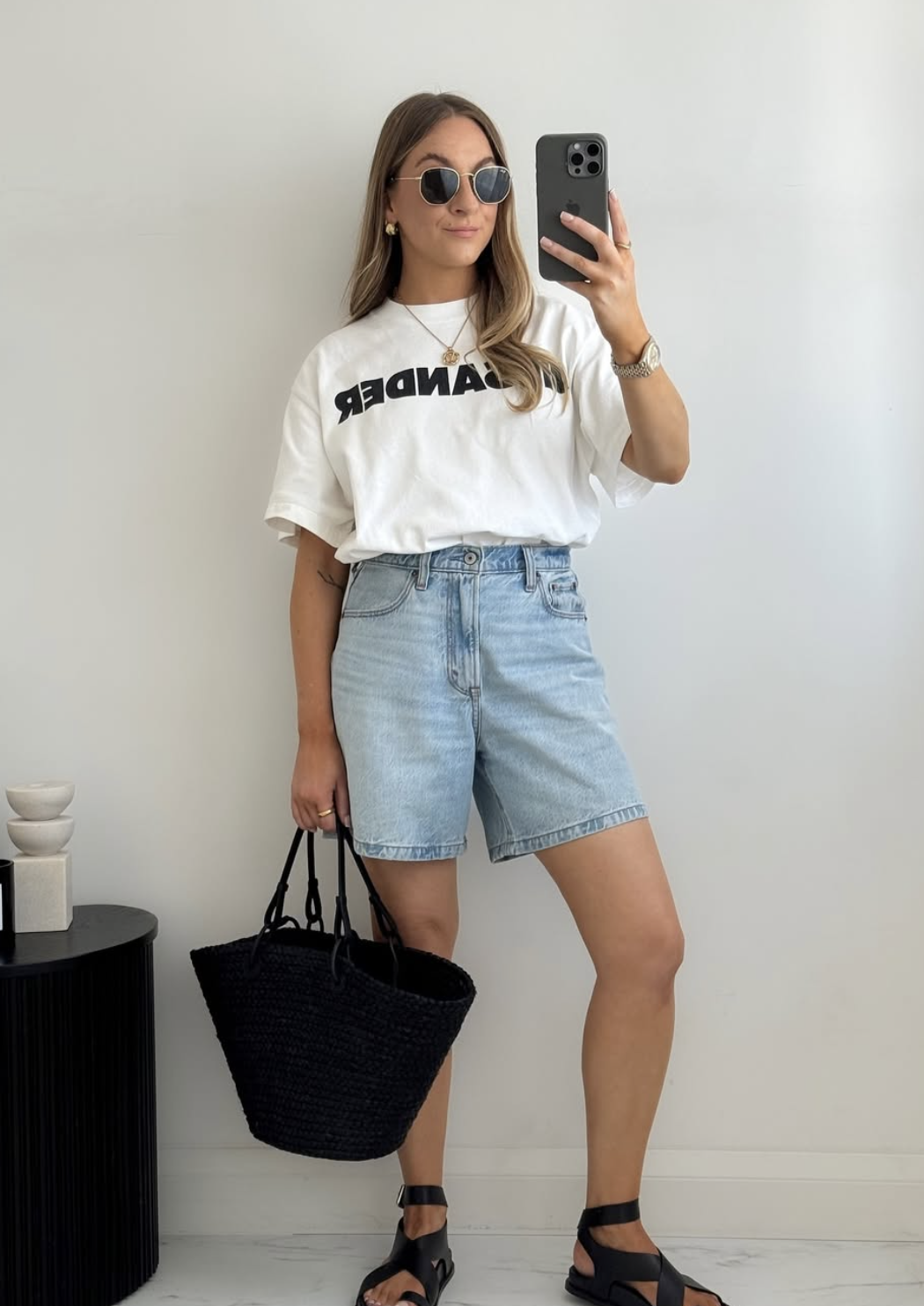Woman wearing a white Wander graphic t-shirt tucked into light blue longline denim shorts with black strappy sandals and a large black woven tote bag.