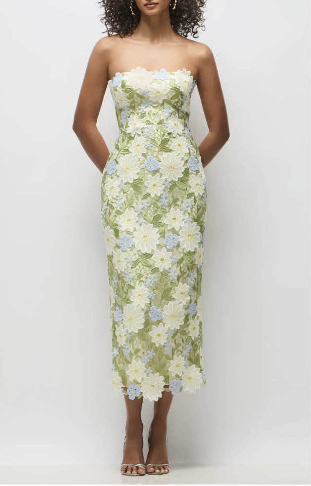 A woman wearing a strapless, tea-length column dress made of intricate 3D floral lace featuring large pale yellow, white, and soft blue daisy motifs, styled with metallic silver strappy heels.