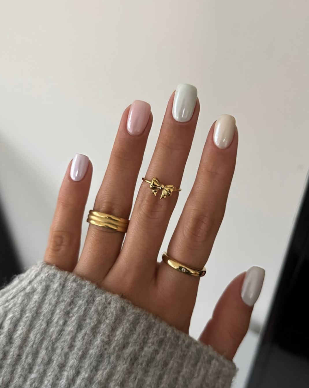 A close-up of a hand wearing a "clean girl" spring manicure with short almond-shaped nails in a glazed, pearlescent pink finish. The look is accessorized with multiple gold rings, including a standout gold bow ring on the index finger.