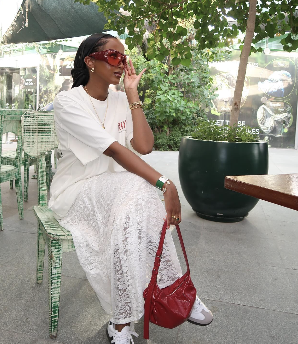 A woman sitting on a green chair wearing a white lace maxi skirt, an oversized white t-shirt, and white sneakers with red accents, holding a red handbag.