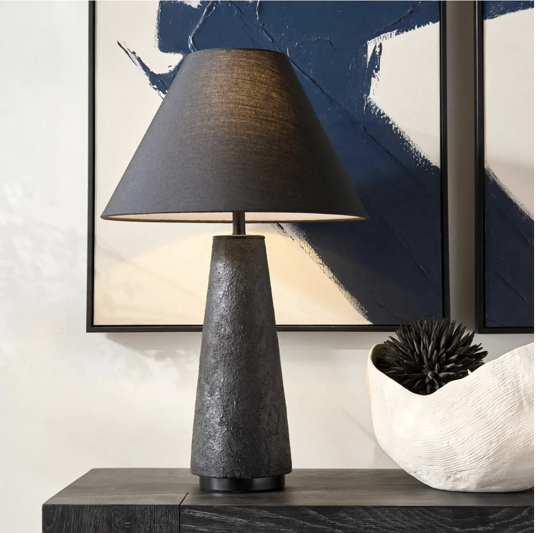 A tall, textured charcoal black ceramic table lamp with a dark drum shade styled on a dark wood console table in front of framed wall art.