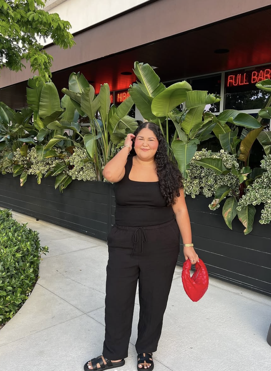 A woman wearing a black tank top and matching wide-leg black linen pants, carrying a bright red woven handbag and standing in front of large tropical banana leaf plants.