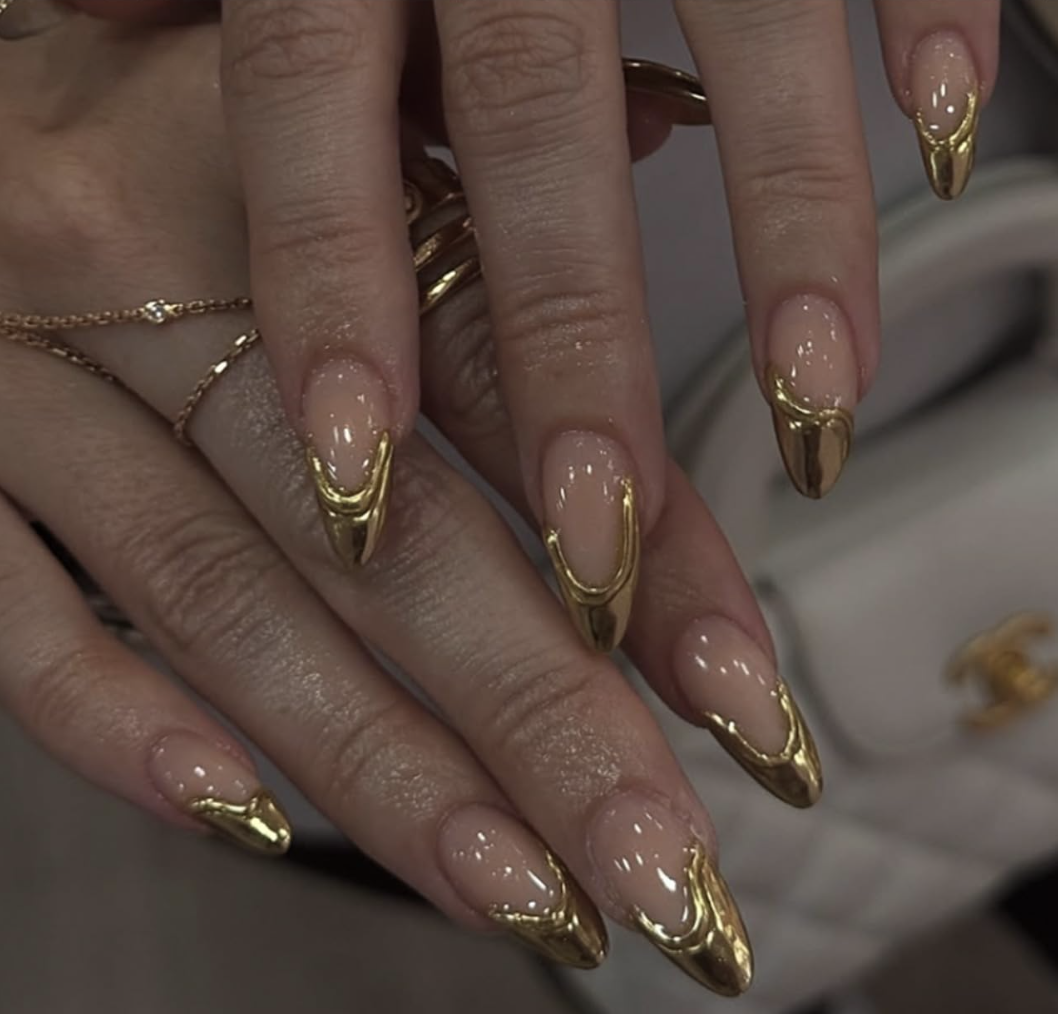 Almond-shaped press-on nails with a nude base, featuring 3D gold chain-link details draped across the tips and shimmering gold glitter accents.