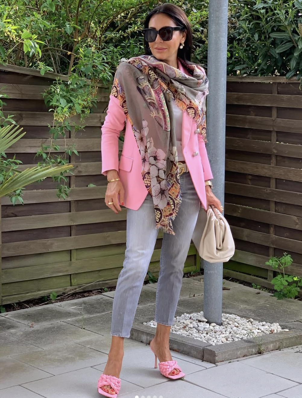 A street style shot of a woman wearing an oversized floral and geometric print scarf draped over a bright pink blazer, paired with grey jeans and a cream clutch bag.