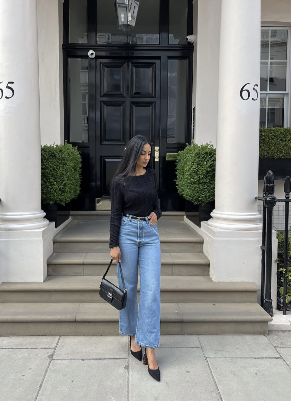 A woman wearing straight-leg light blue jeans paired with a black long-sleeve bodysuit, a thin black leather belt, a black shoulder bag, and pointed-toe black heels.
