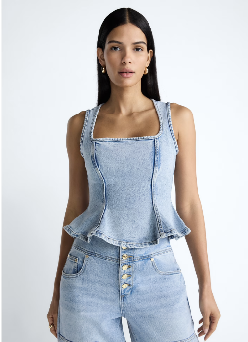 A woman wearing a light-wash denim peplum top with thick straps and a structured bodice from the Scoop x Devil Wears Prada collection, styled with matching jeans.