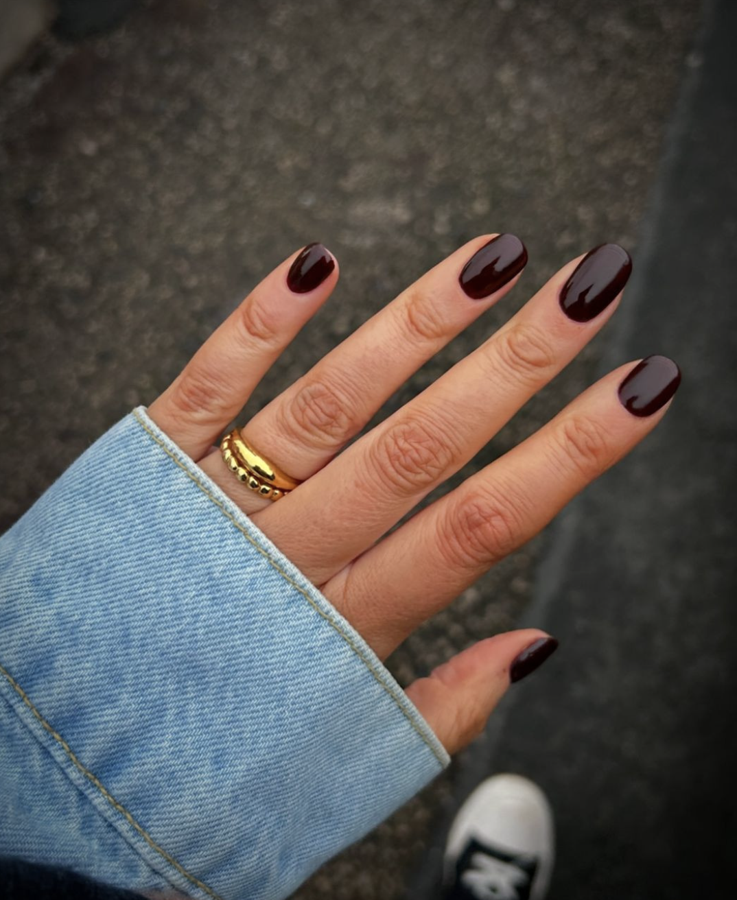 Close-up of short nails painted in a glossy, dark black-cherry nail polish paired with a denim jacket and gold rings.