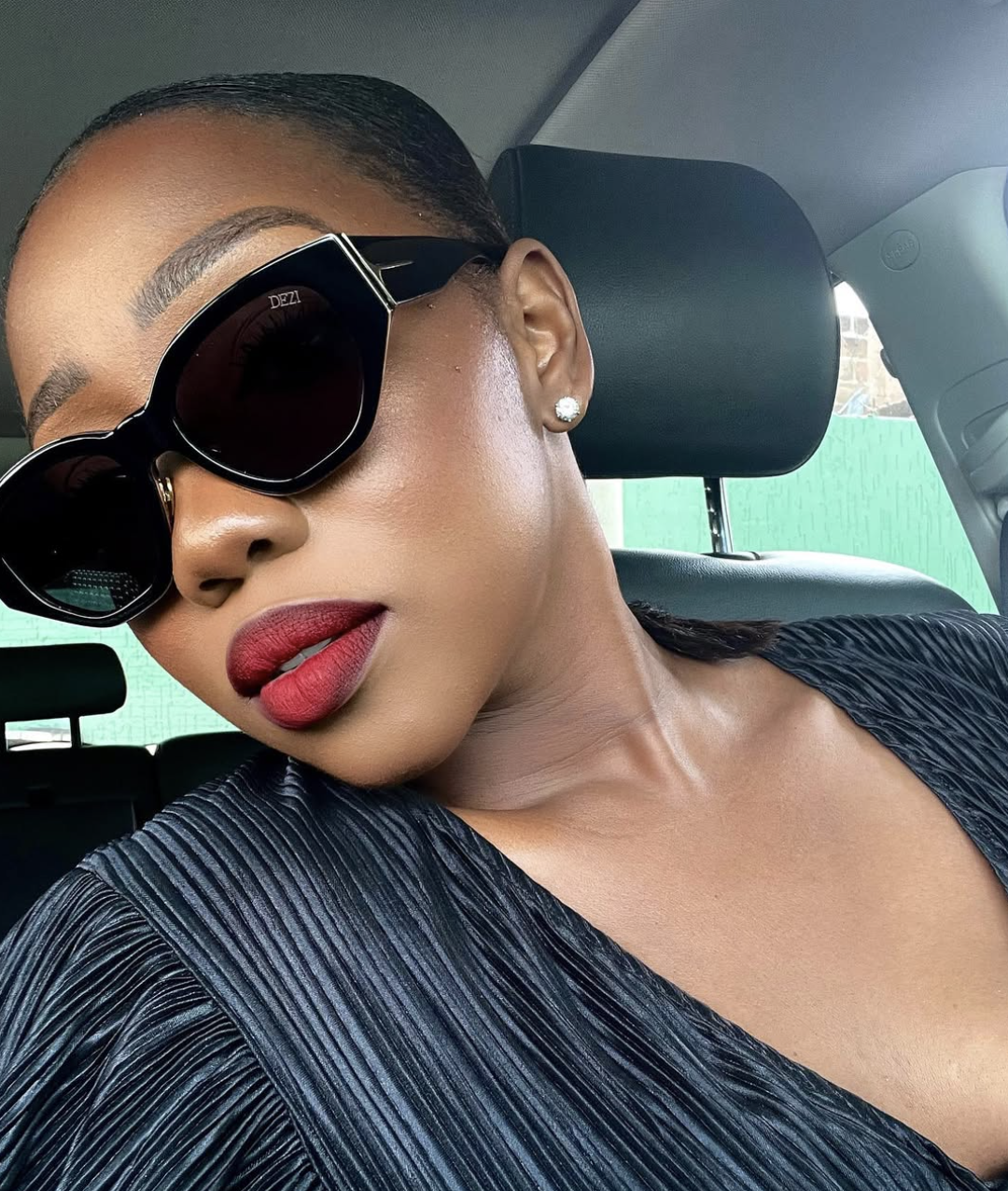 A close-up of a woman with deep skin tone showcasing a Winter color season look, wearing bold red lip liner with a lighter red gloss and oversized black square sunglasses with a metallic gold top rim.