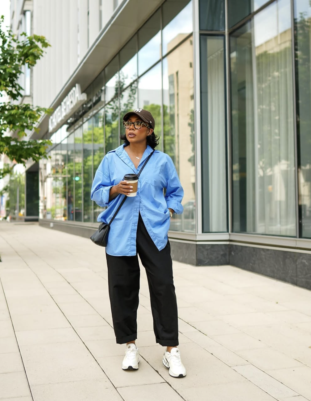 : A woman holding a coffee cup, wearing an oversized bright blue button-down shirt over relaxed black trousers, finished with white technical sneakers and a black crossbody bag.
