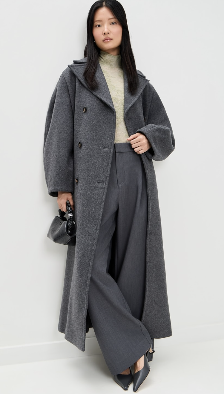 A person wearing a long, charcoal grey wool trench coat with a double-breasted design, paired with matching wide-leg trousers and black pointed-toe heels.