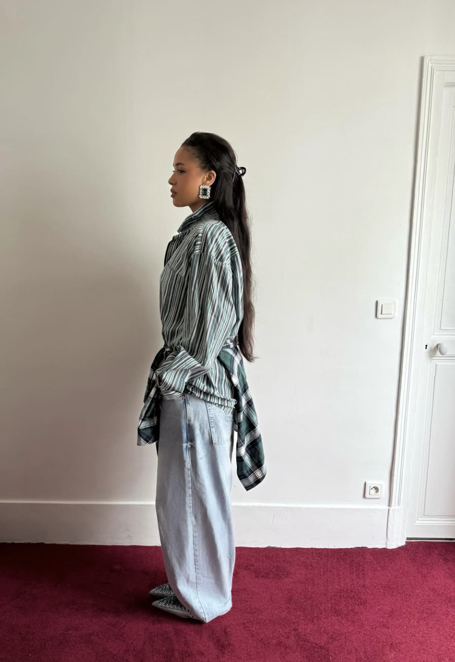 A woman wearing light-wash baggy jeans with a striped green and white relaxed button-down shirt half-tucked to show the waistline and highlight proper proportions.