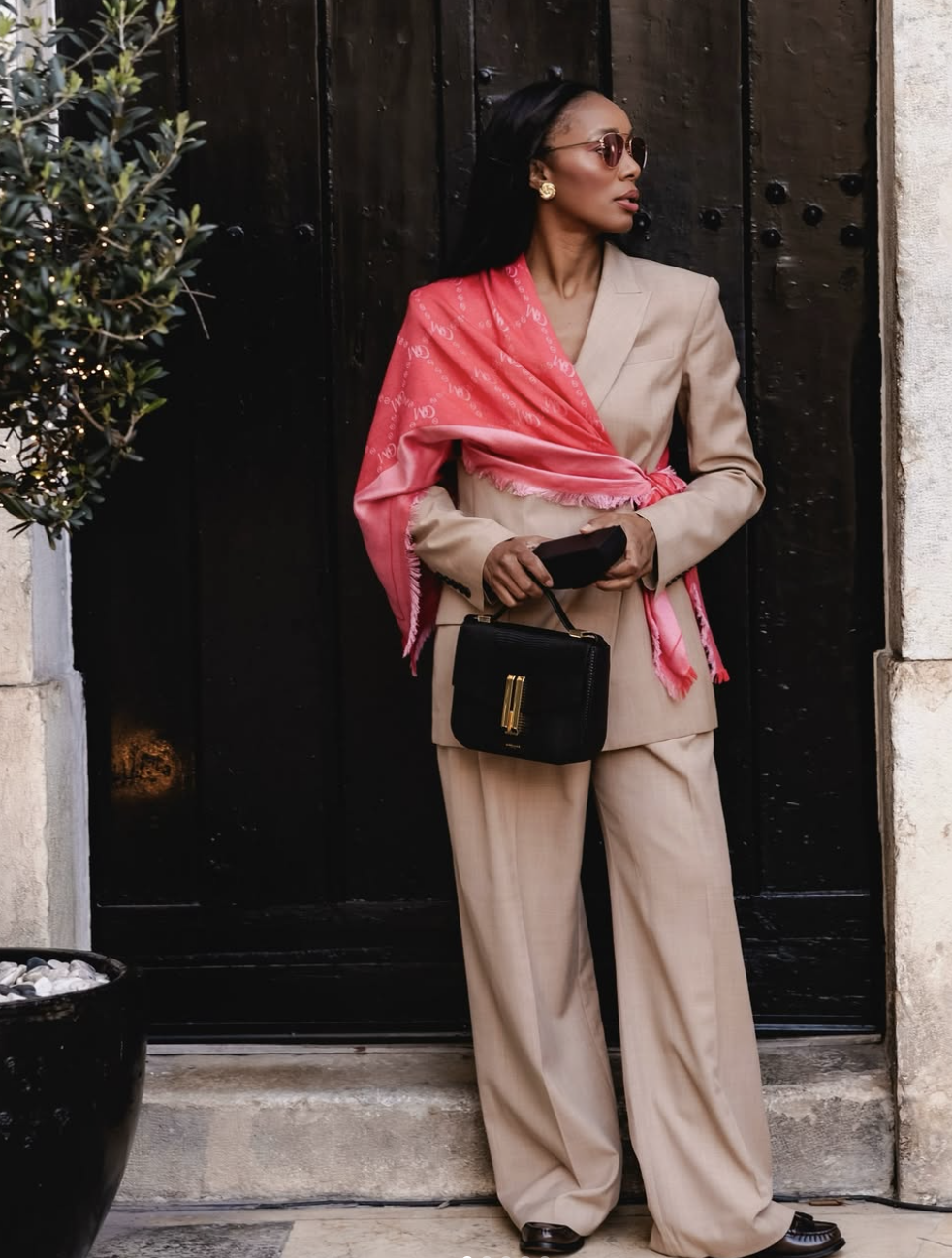 A close-up street style shot of a woman wearing an oversized pink silk statement scarf draped over a tailored beige pantsuit, holding a structured black leather handbag.