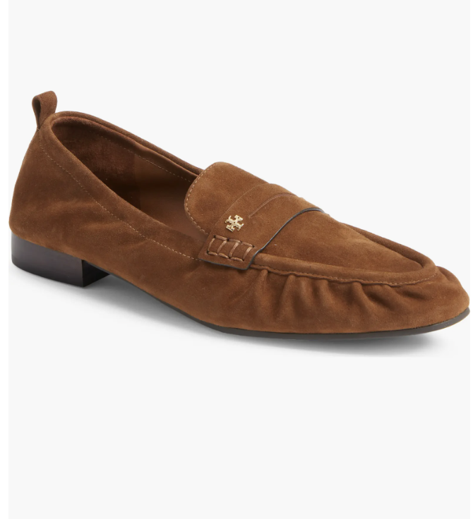 A pair of cognac brown suede loafers featuring a low block heel, moc-toe stitching, and a gold emblem detail.