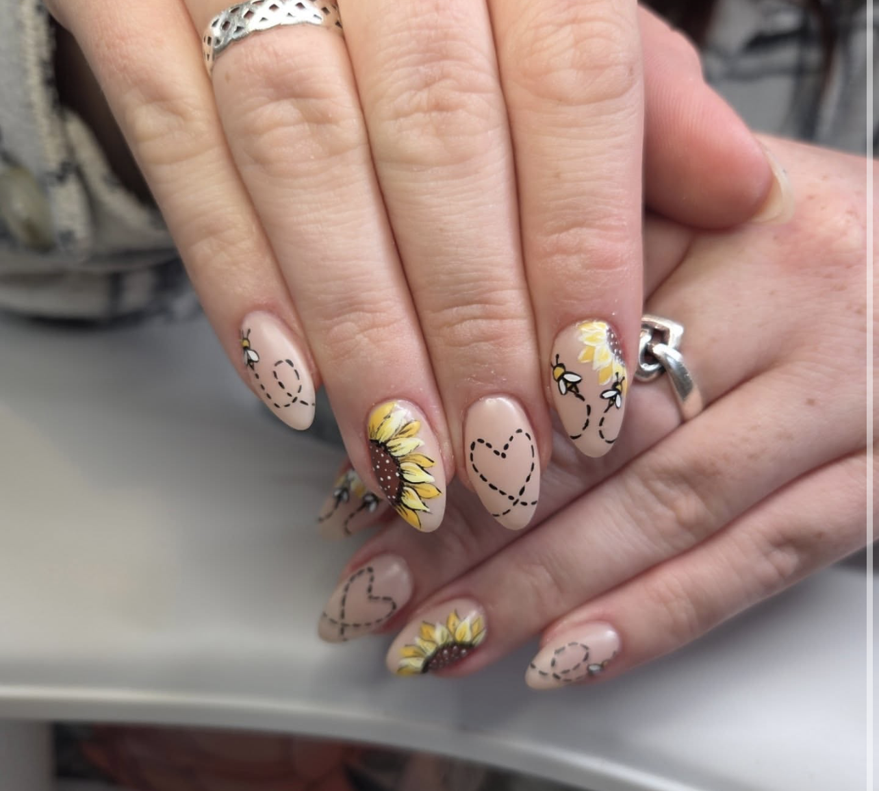 Almond-shaped nude nails with hand-painted sunflowers and a tiny honeybee leaving a dotted heart-shaped flight trail.