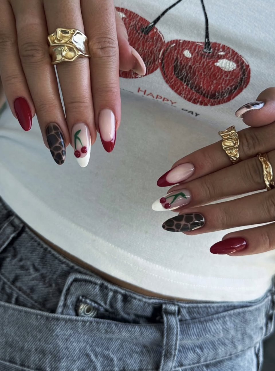 A close-up of almond-shaped nails featuring a mix of glossy cherry red polish, tortoiseshell print accent nails, and a white French tip with hand-painted cherry details.