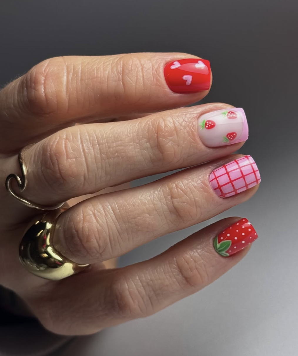 A close-up of a playful summer manicure featuring bright red nails with white hearts, pink gingham check patterns, and a white accent nail with hand-painted strawberries, highlighting cute June nail ideas.