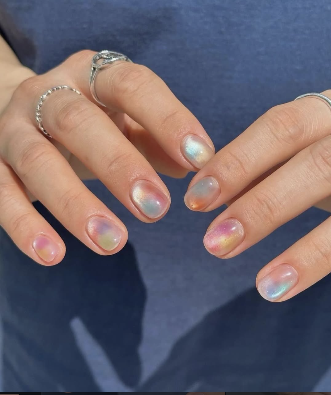 Short almond-shaped nails featuring a hazy aura effect with blended pastel shades of pink, blue, and yellow on a sheer, shimmering base.