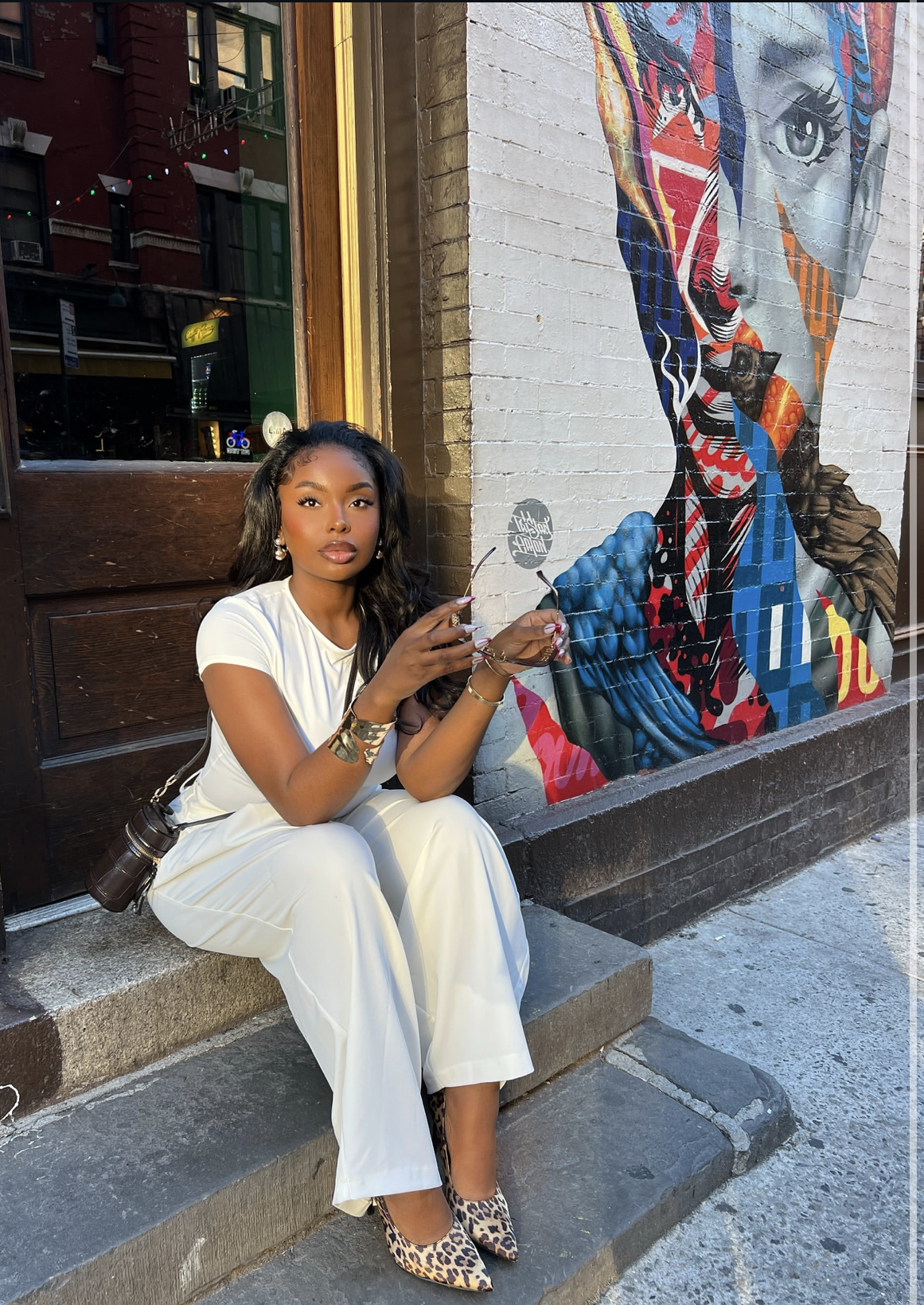 A woman sitting on stone steps wearing a white t-shirt and wide-leg white linen pants, paired with leopard print pointed-toe mules and layered gold jewelry.