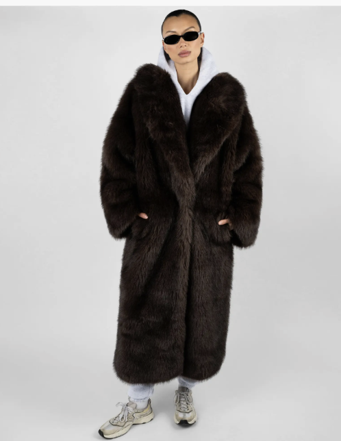 A woman wearing a long dark brown faux fur jacket over a white hoodie, demonstrating how to use a long coat as a "third piece" to streamline a baggy jeans silhouette.