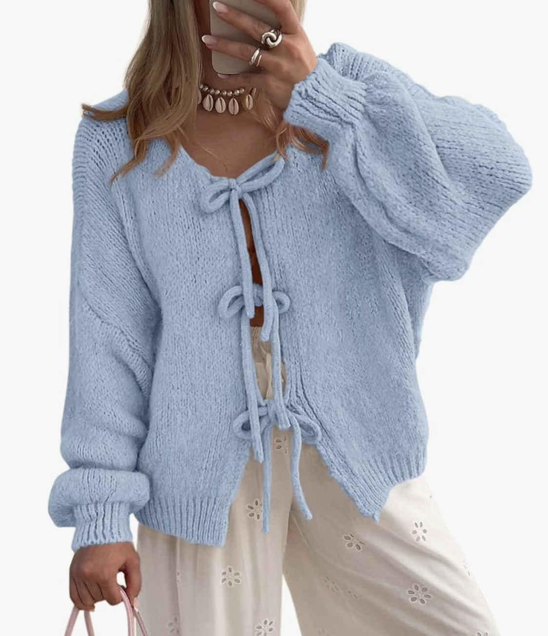 A light blue chunky knit cardigan featuring three delicate black bow tie-front closures, styled with cream-colored floral trousers.