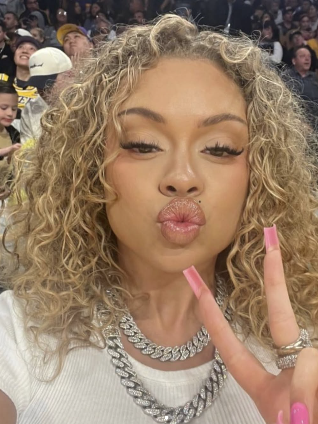 Latto showcasing a voluminous warm platinum blonde curly hairstyle while wearing layered silver chain necklaces.