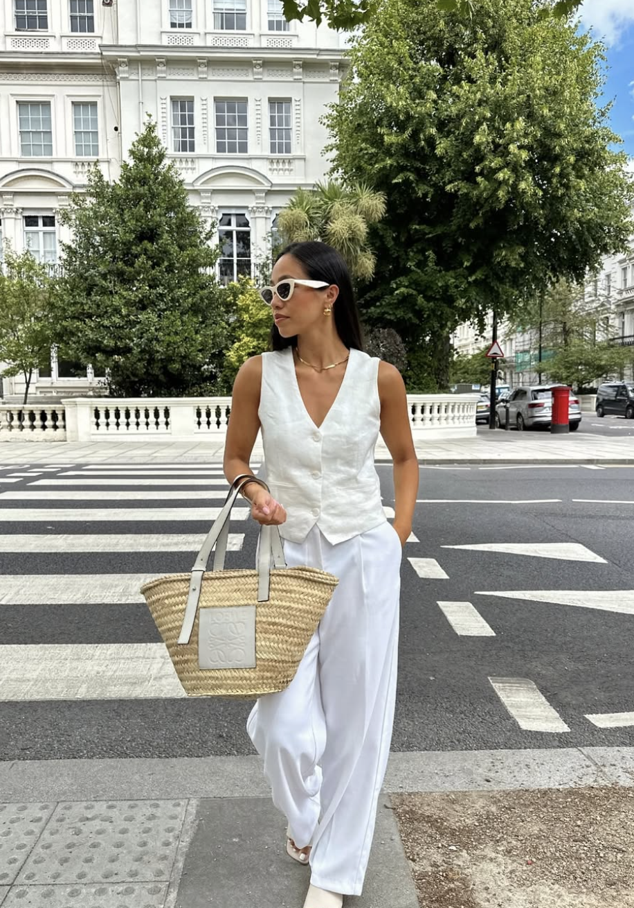 A woman wearing a tailored oatmeal-colored linen vest with matching high-waisted wide-leg linen trousers, carrying a large straw tote bag while standing on a city sidewalk.
