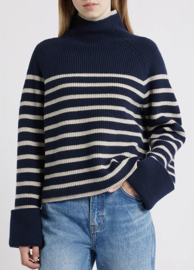 Woman wearing a navy and cream striped mock-neck sweater with wide cuffs and light-wash jeans.