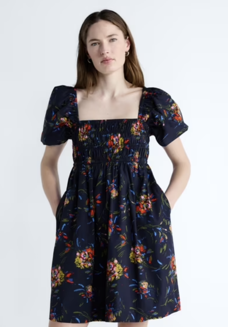 A product shot of a woman wearing a high-waisted dark navy blue midi dress featuring a vibrant multi-colored floral pattern, classic cap sleeves, a square neckline, and practical side pockets.