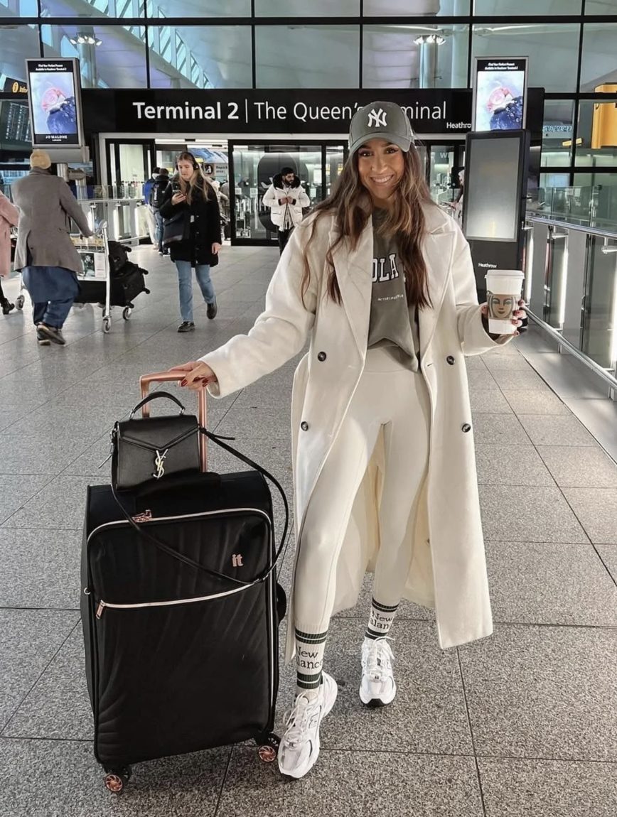 Long cream coat over a muted graphic hoodie and pale leggings, with patterned socks, white sneakers and a dark baseball cap.