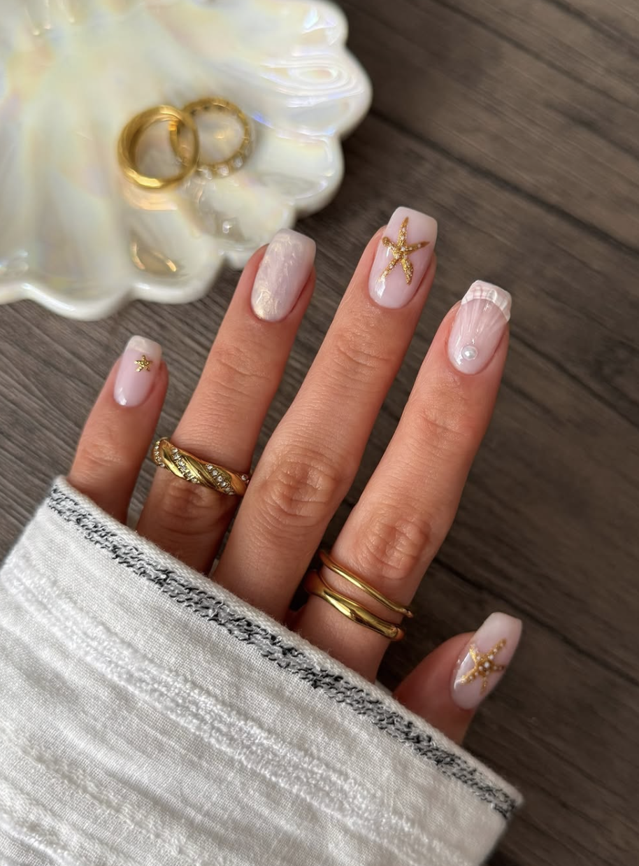 A close-up of long, square-shaped nails with a classic white French manicure, accessorized with tiny 3D gold starfish charms, a single pearl embellishment, and chunky gold rings.