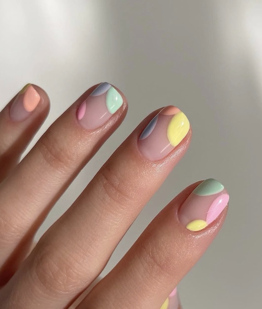 A close-up of short round nails with a clear base featuring organic, pebble-like abstract shapes in pastel peach, mint green, lavender, and sunny yellow, mimicking the pattern of dyed Easter eggs.