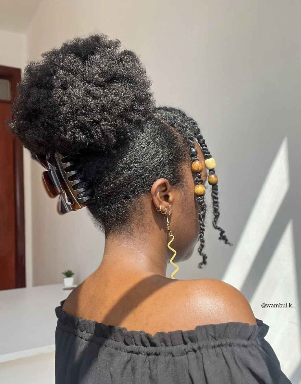 Back view of a woman with braided hair styled in an updo secured with a tortoiseshell claw clip, wearing a black off-the-shoulder top and gold drop earrings.