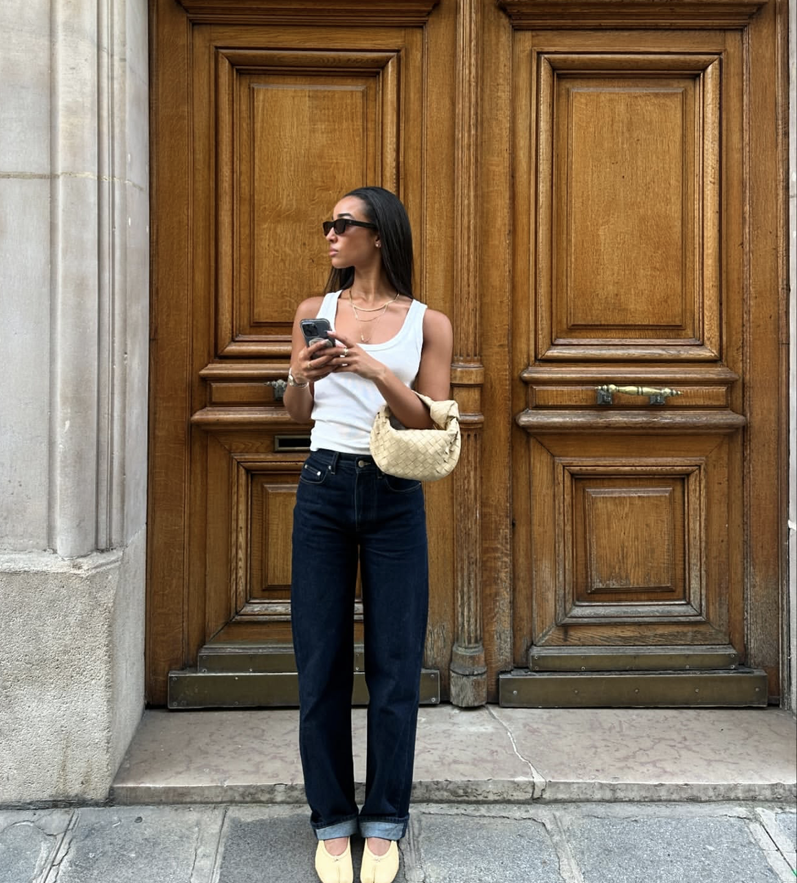 Front view of a woman wearing a white ribbed tank top tucked into high-waisted dark wash jeans with a cuffed hem, paired with beige flat shoes.