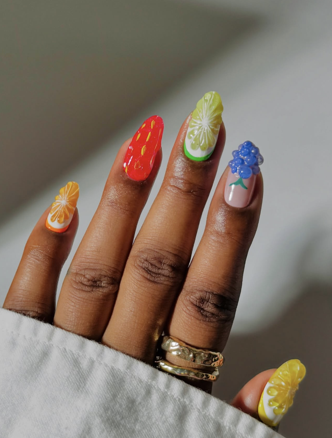 Close-up of a hand with long nails showcasing a vibrant summer fruit salad manicure featuring detailed, 3D-textured nail art of sliced oranges, strawberries, kiwis, and grapes on a sheer base.