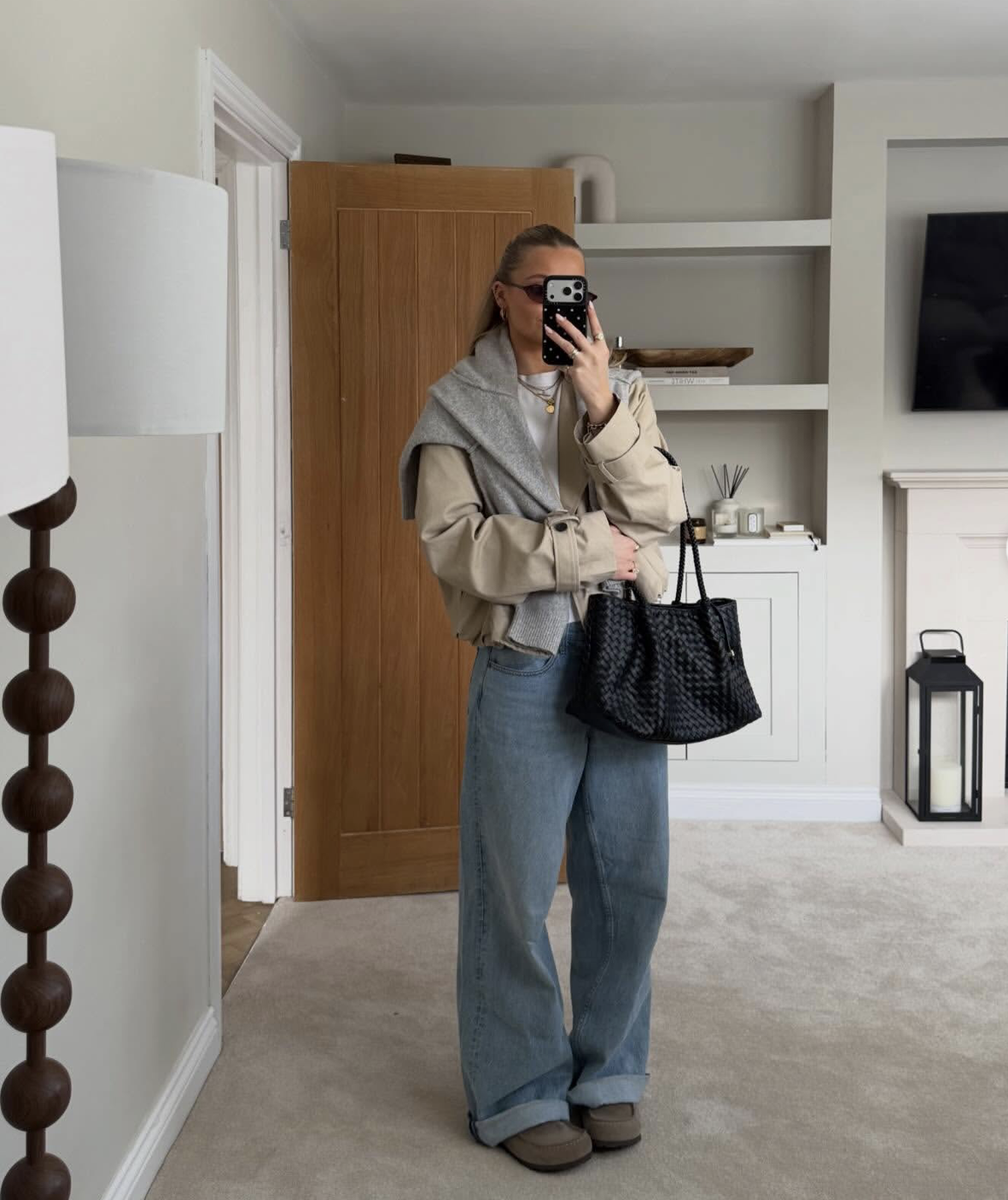 Digital creator Lissie Judd taking a mirror selfie wearing an oversized tan jacket, a grey sweater draped over her shoulders, and light-wash wide-leg jeans with a black woven tote bag.